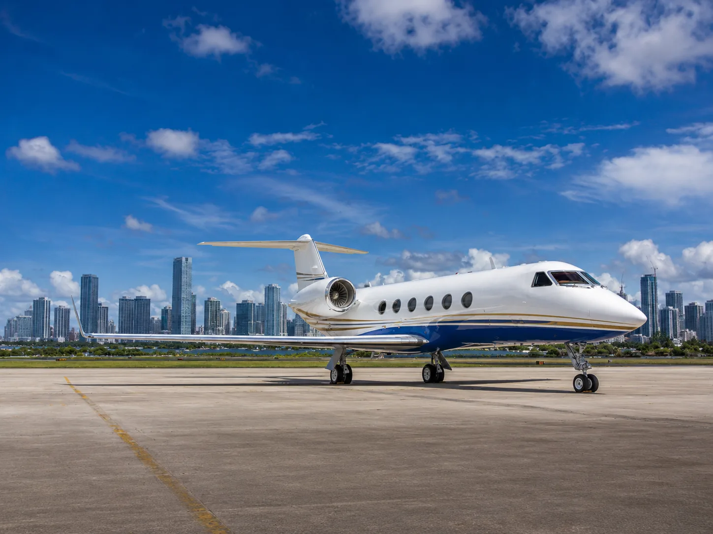 A white and blue Gulfstream business jet is parked on an airport tarmac with a city skyline under a blue sky.