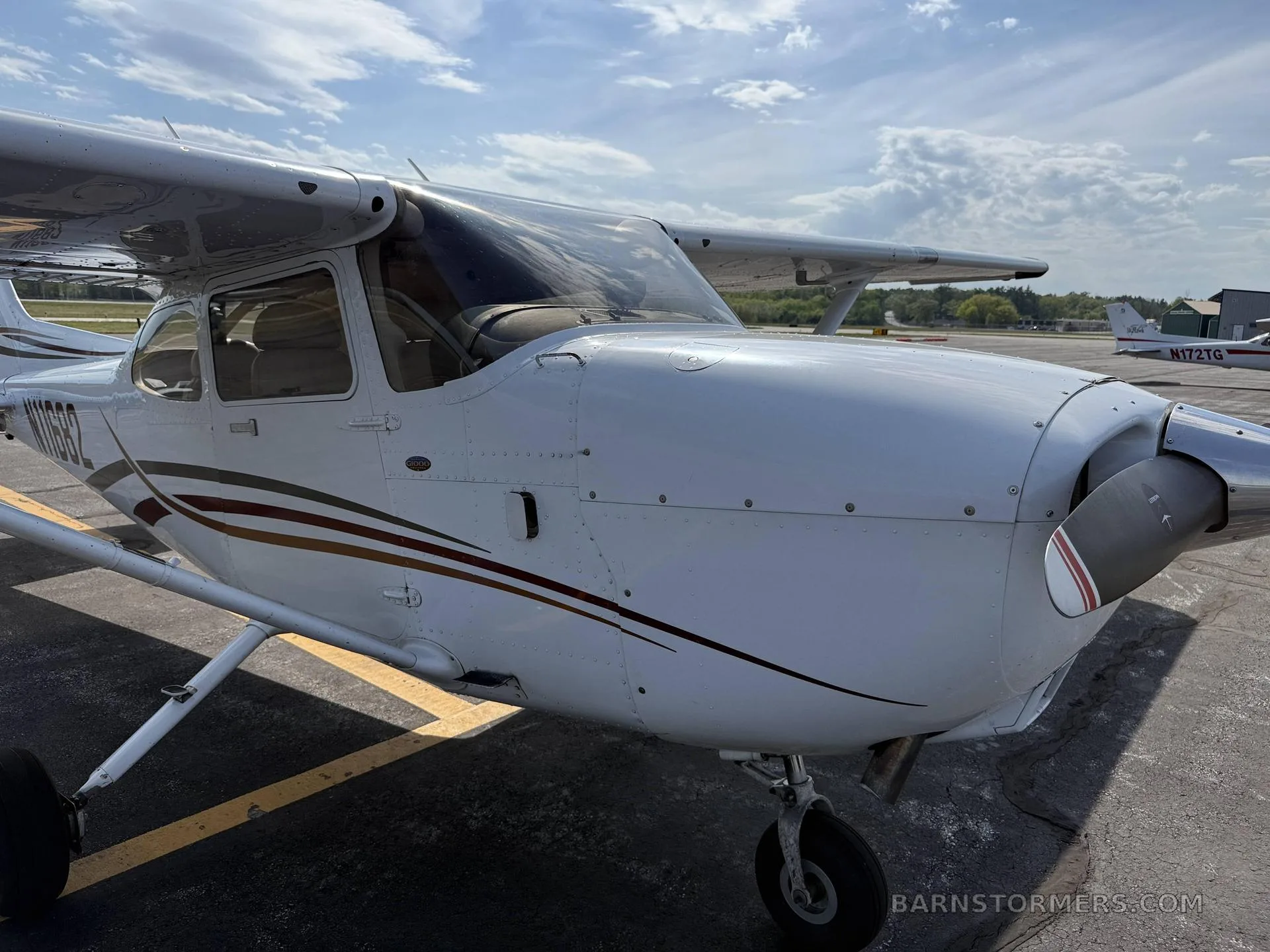 A close-up front-right view of a white Cessna 172 aircraft with brown and black stripes, parked on an airfield.
