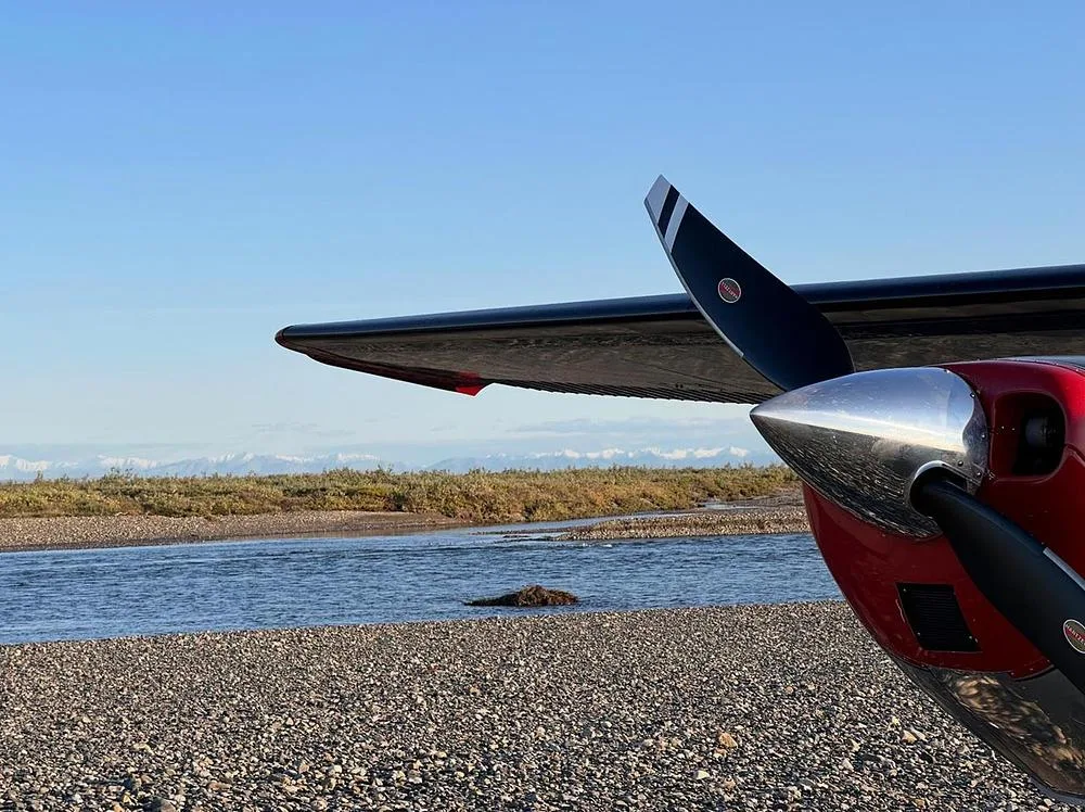 A close-up view of the propeller and wing of a silver and red aircraft on a gravel bar by a river.