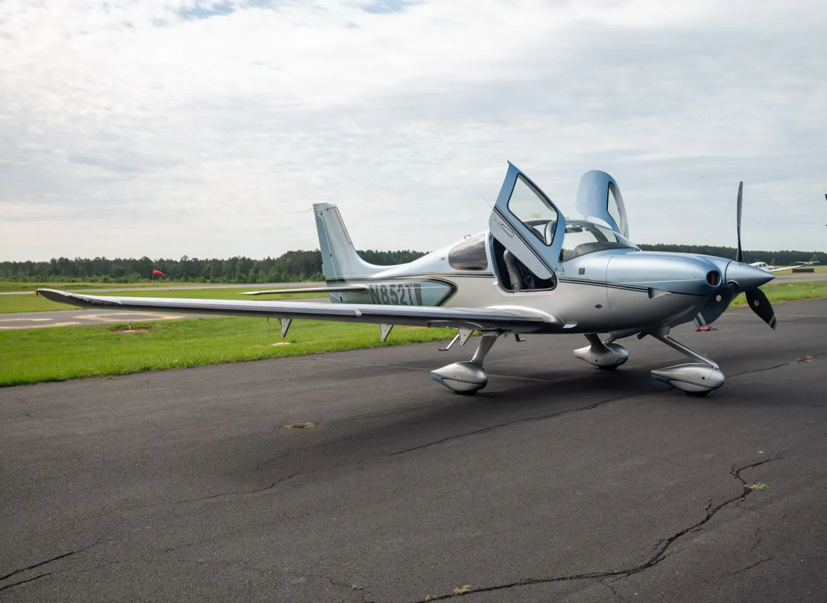 A side view of a blue and silver Cirrus SR22 aircraft, N852TW, with its gull-wing doors open on an asphalt tarmac.