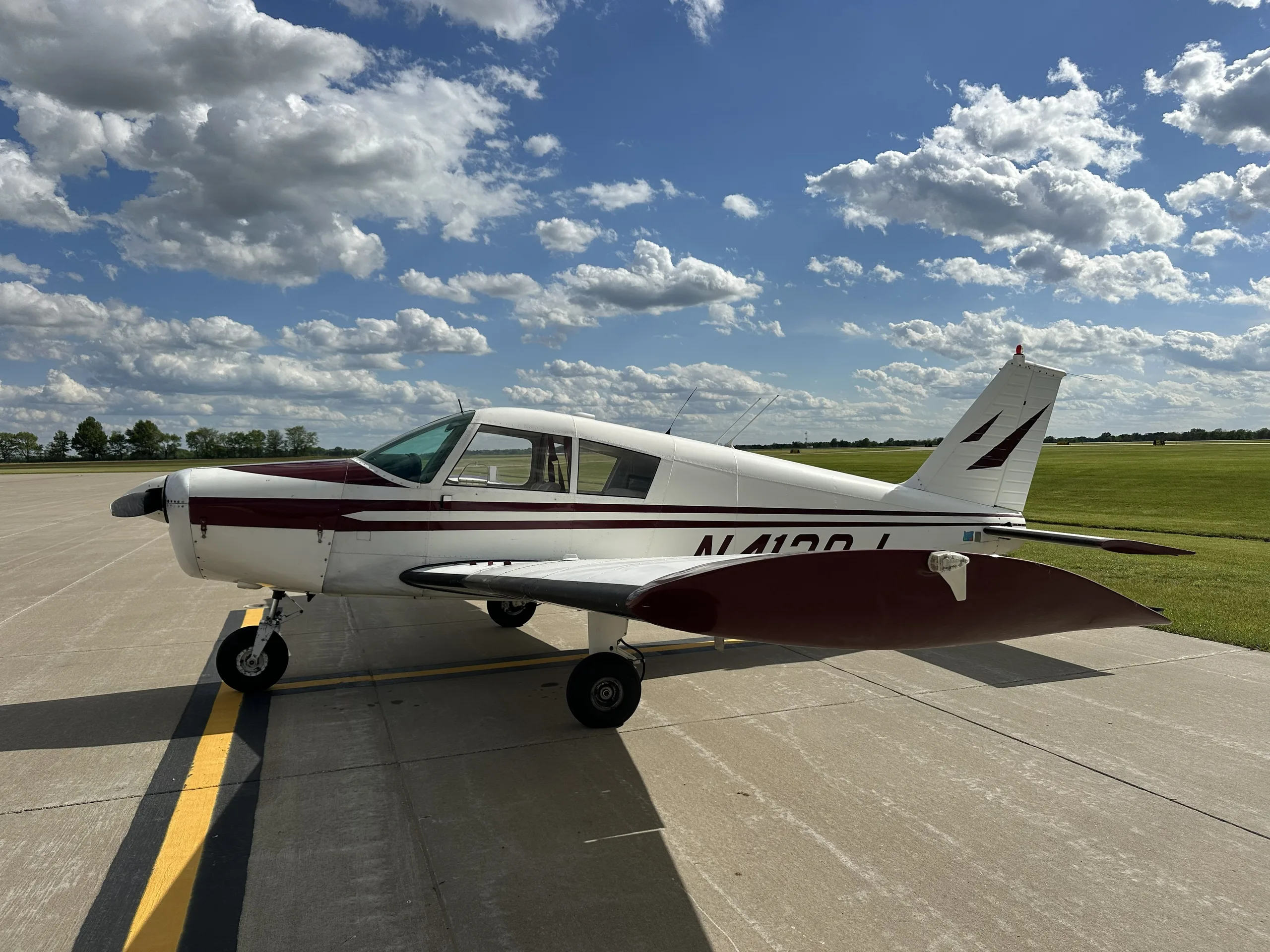A side view of a white and maroon single-engine aircraft, likely a Piper Cherokee, parked on an asphalt ramp with green grass in the background.