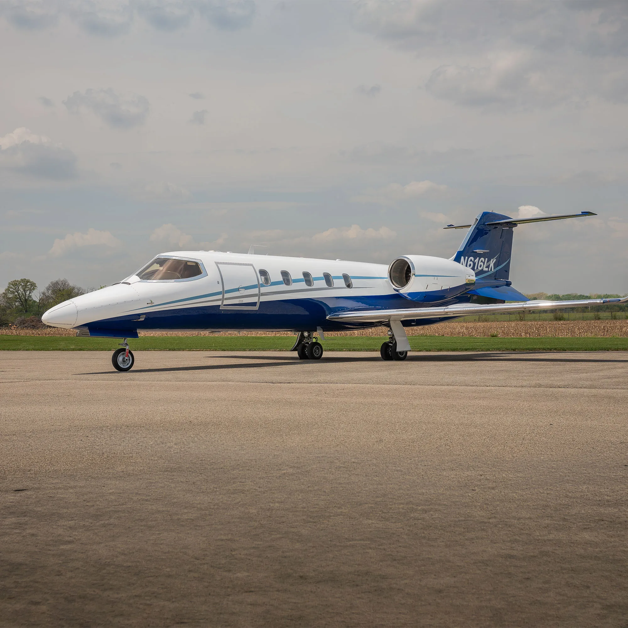 A white and blue Learjet N616LK parked on an airfield under a cloudy sky.