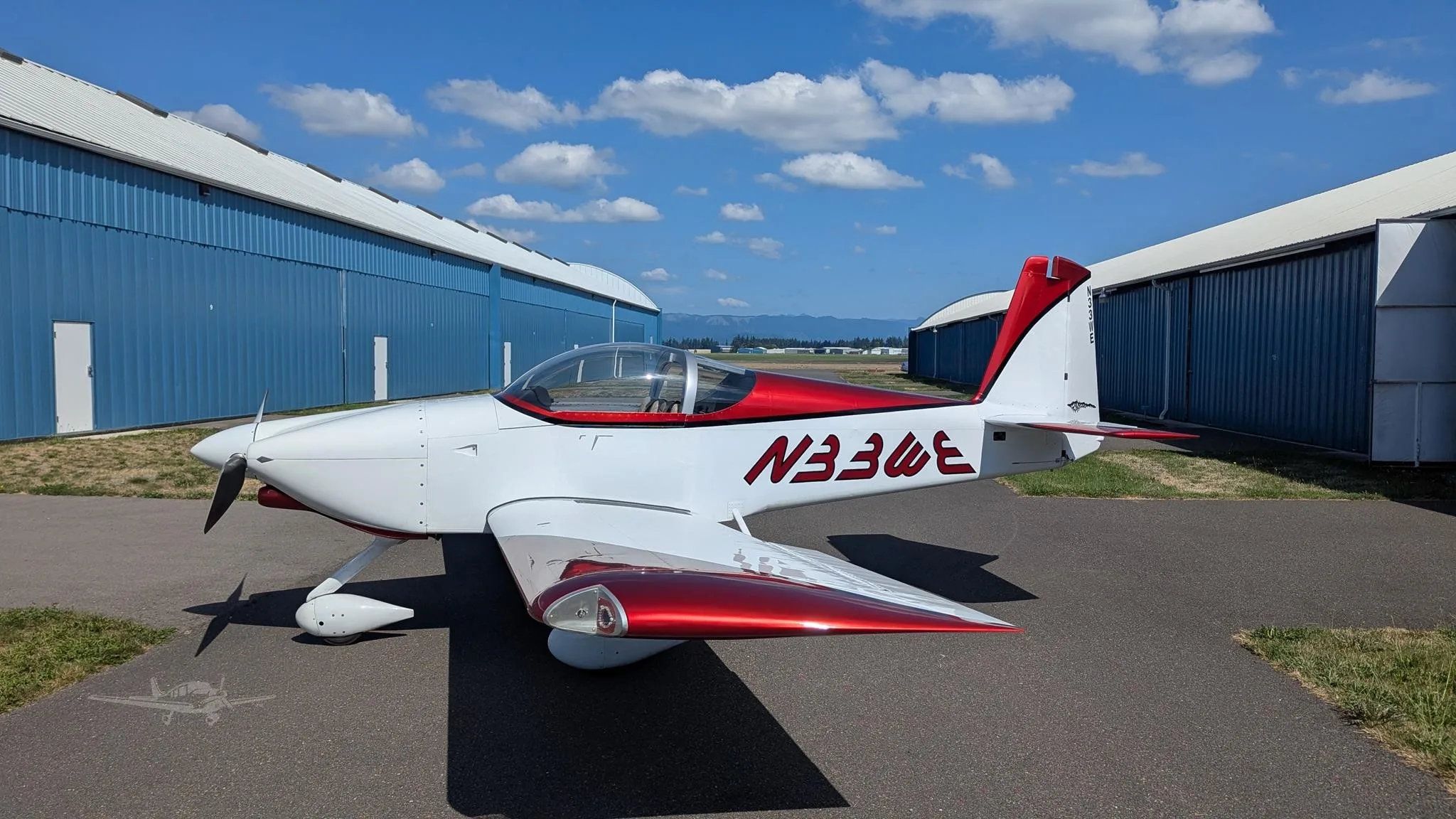 A white and metallic red single-engine aircraft, registration N33WE, parked on an airfield tarmac in front of blue hangars.