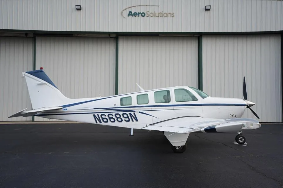 A white Beechcraft Bonanza with blue and gray stripes, registration N6689N, parked on an asphalt ramp in front of a hangar.