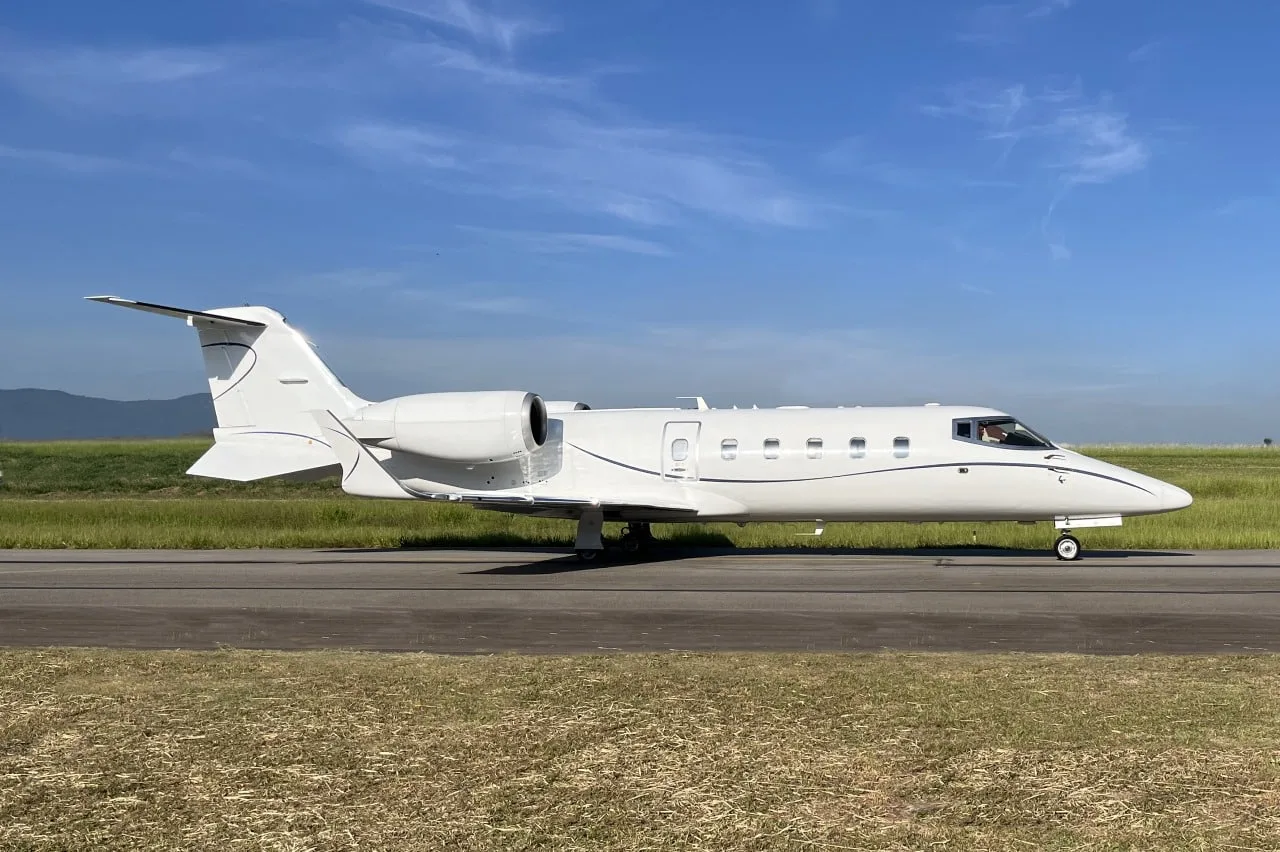 A white business jet with blue pinstripes is parked on a runway under a clear blue sky, viewed from the side.
