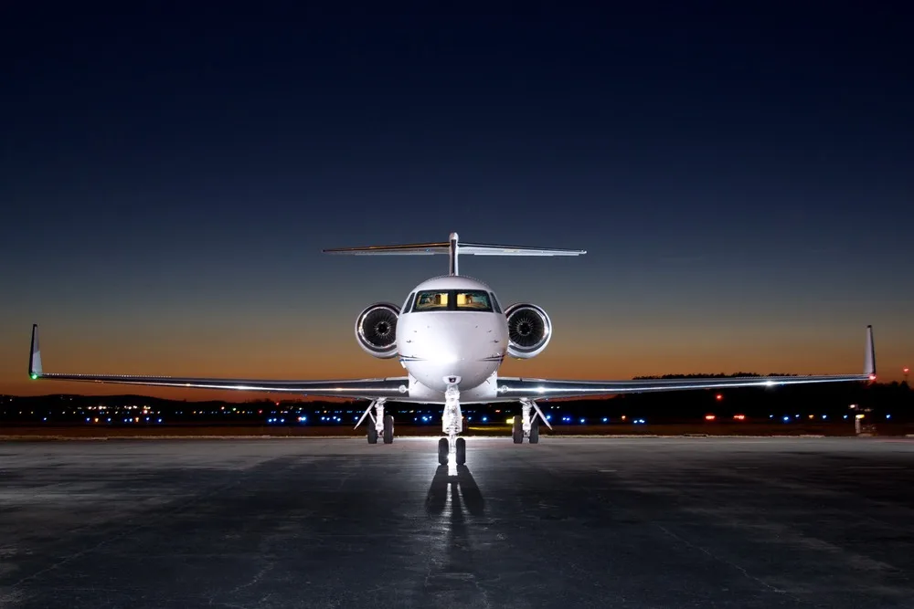 A white Gulfstream G450 business jet with a blue stripe on the tarmac at dusk, viewed head-on.