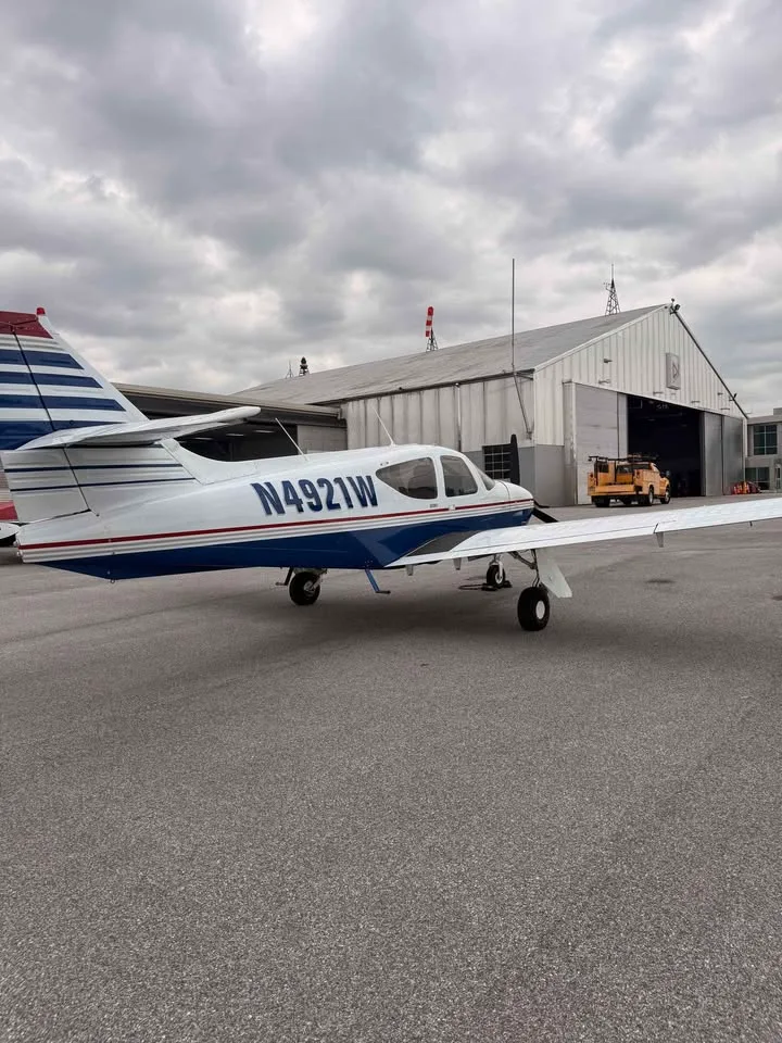A rear-left view of a white and blue single-engine aircraft with registration N4921W parked on the tarmac next to a hangar.
