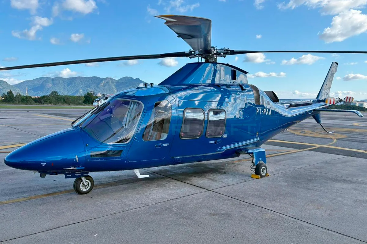 A blue Leonardo AW109 helicopter (PT-PAA) parked on an airfield with mountains in the background.