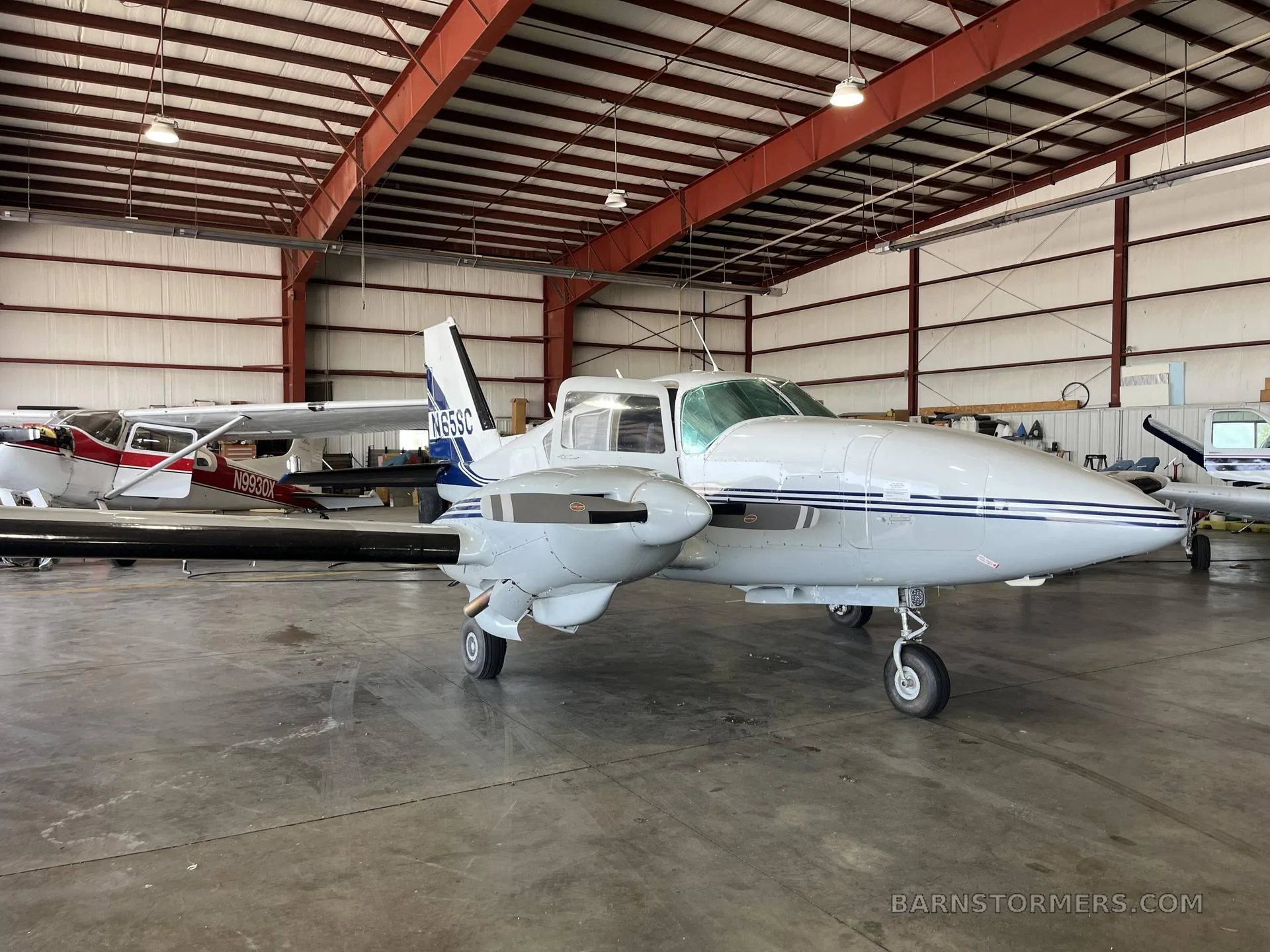 A white twin-engine aircraft with blue stripes (N65SC) parked in a hangar, with another aircraft partially visible.