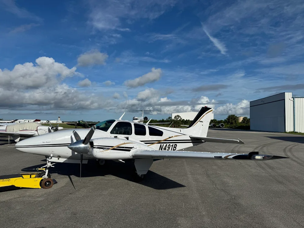 A white twin-engine Beechcraft Baron aircraft with black and gold stripes, registration N491B, parked on an airfield next to a hangar.