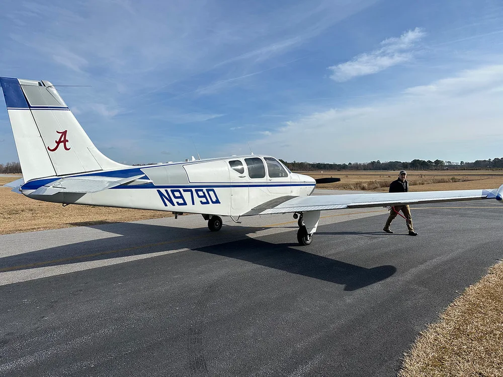 A white and blue Beechcraft Debonair N979Q with a person walking by its wing on a sunny day at an airfield.