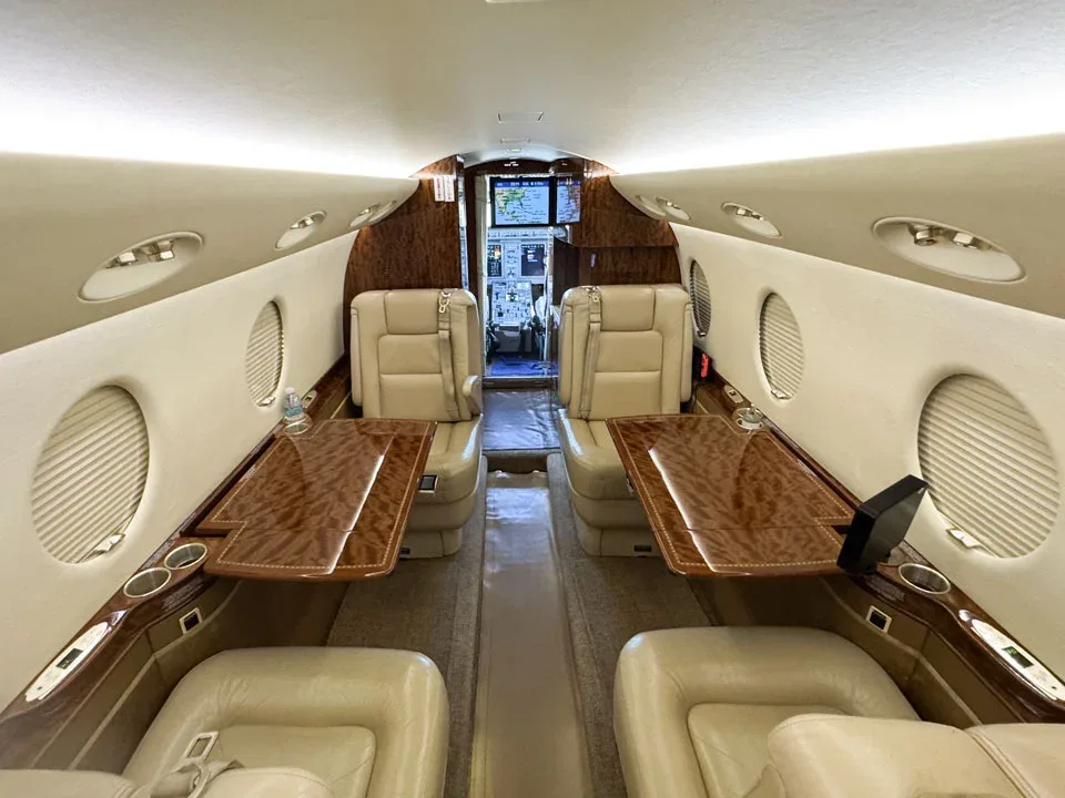 The interior cabin of a private jet with beige seats, wood tables, and a view into the cockpit.