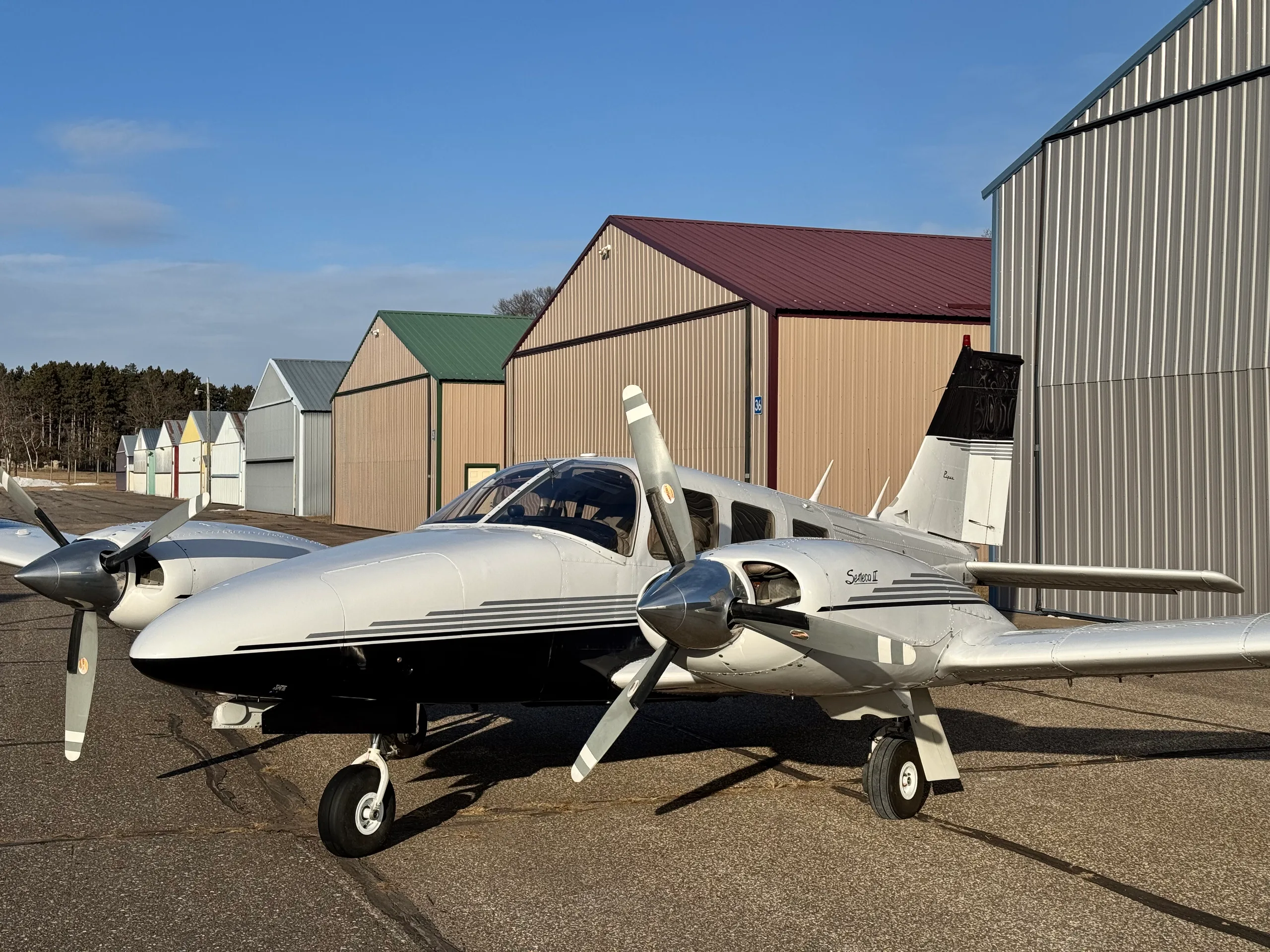 A white and black Piper Seneca II twin-engine aircraft with grey stripes is parked on an asphalt ramp in front of hangars under a clear blue sky.
