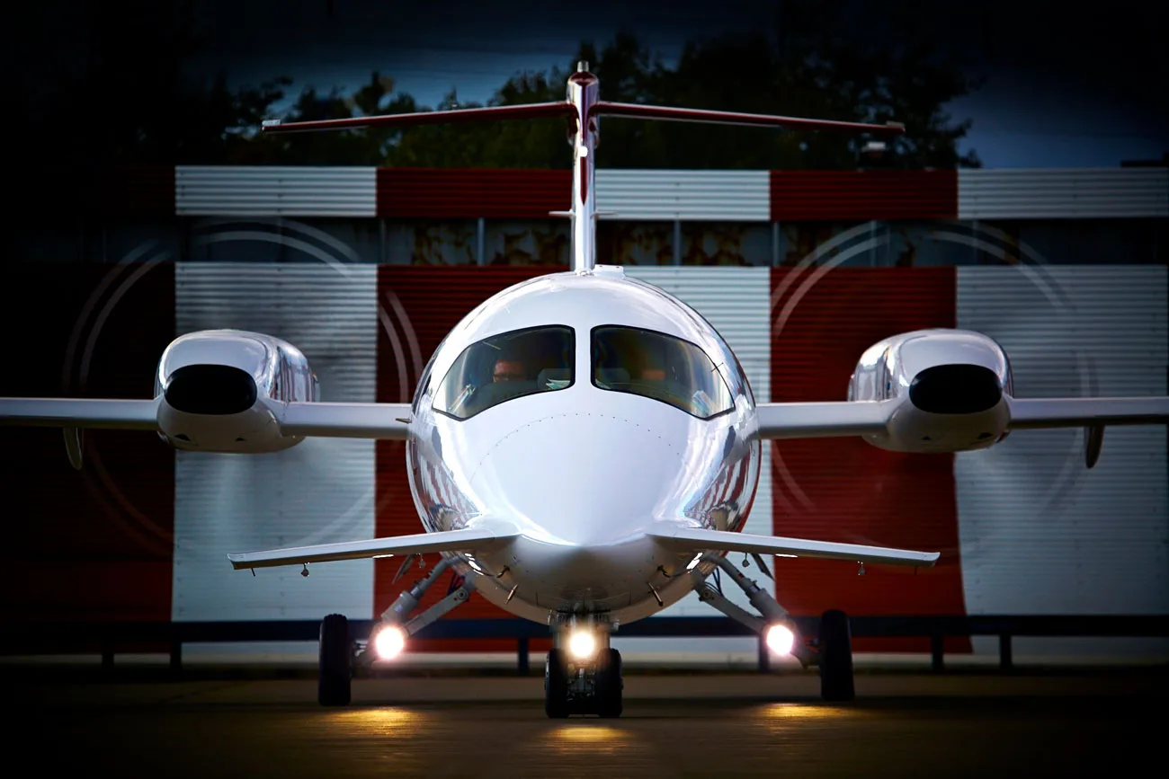 Front view of a white Piaggio P.180 Avanti twin-turboprop aircraft with landing lights on, parked in front of a red and white striped building.