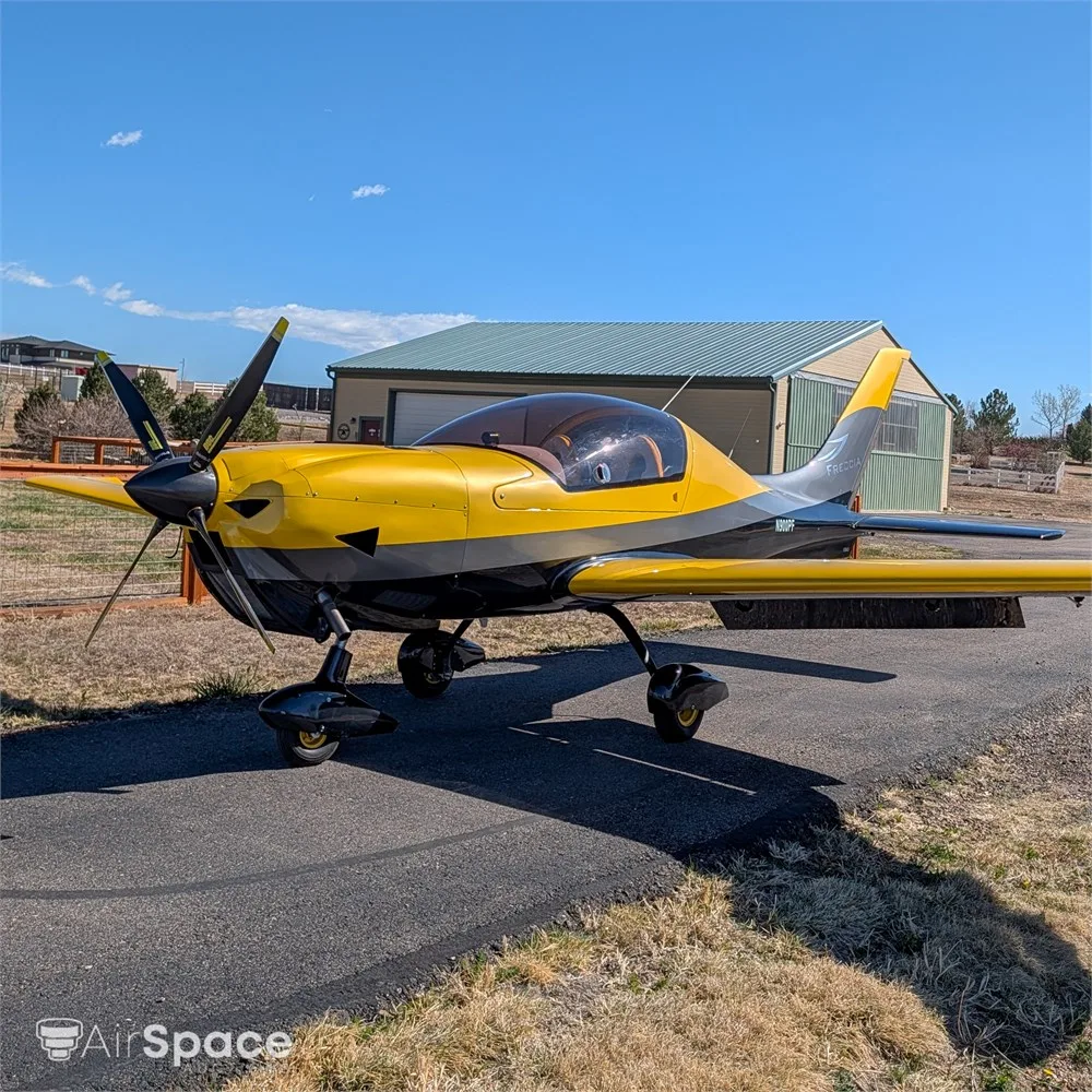 A side view of a yellow and black Freccia F200 aircraft with N900PF on the fuselage, parked on an asphalt ramp in front of a hangar.