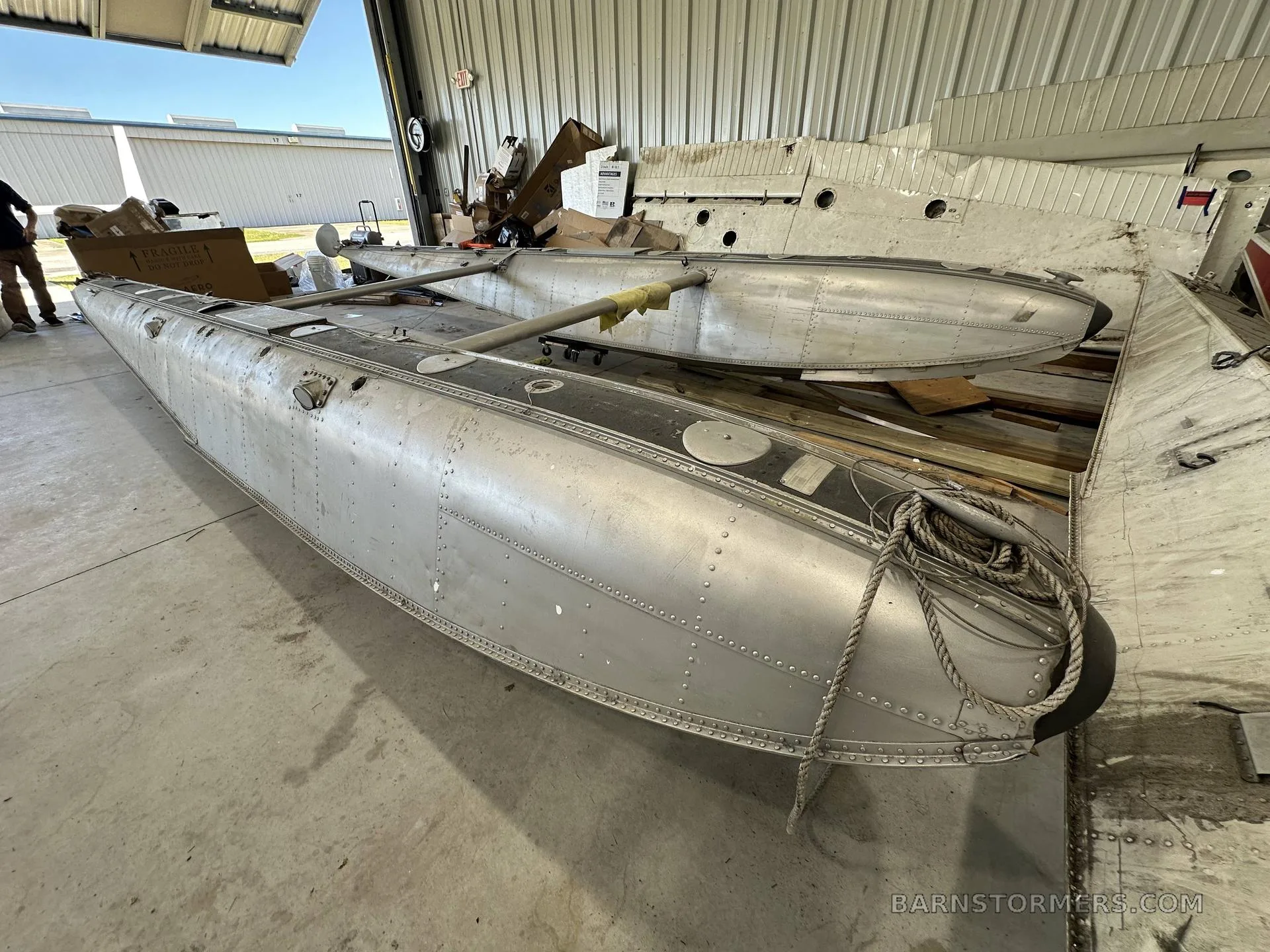 Two large aluminum aircraft floats are stored in a hangar, with one float partially disassembled.