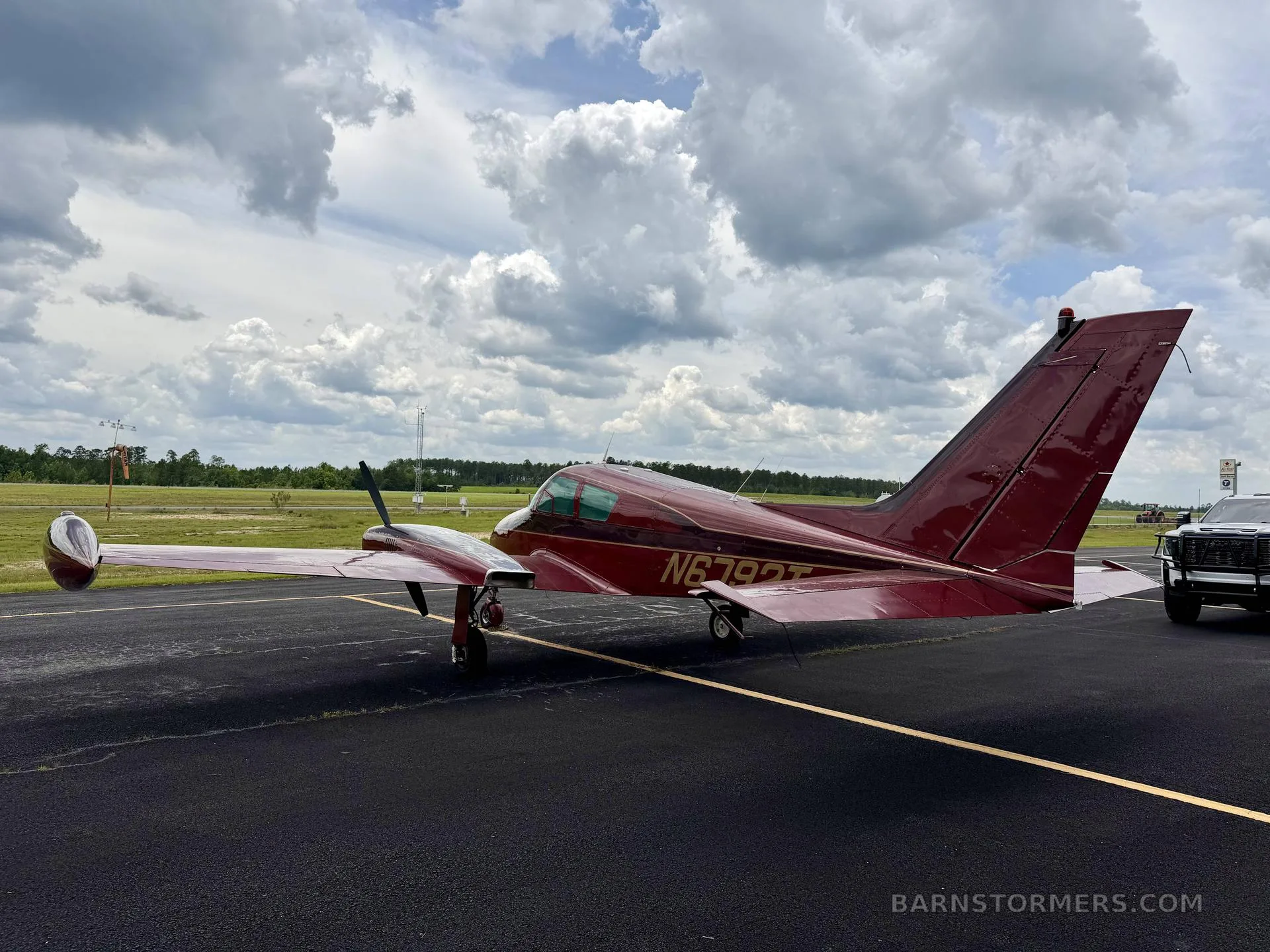 A rear-quarter view of a red twin-engine aircraft, likely a Beechcraft Baron, with registration N6792T, parked on an asphalt runway with a distant field.