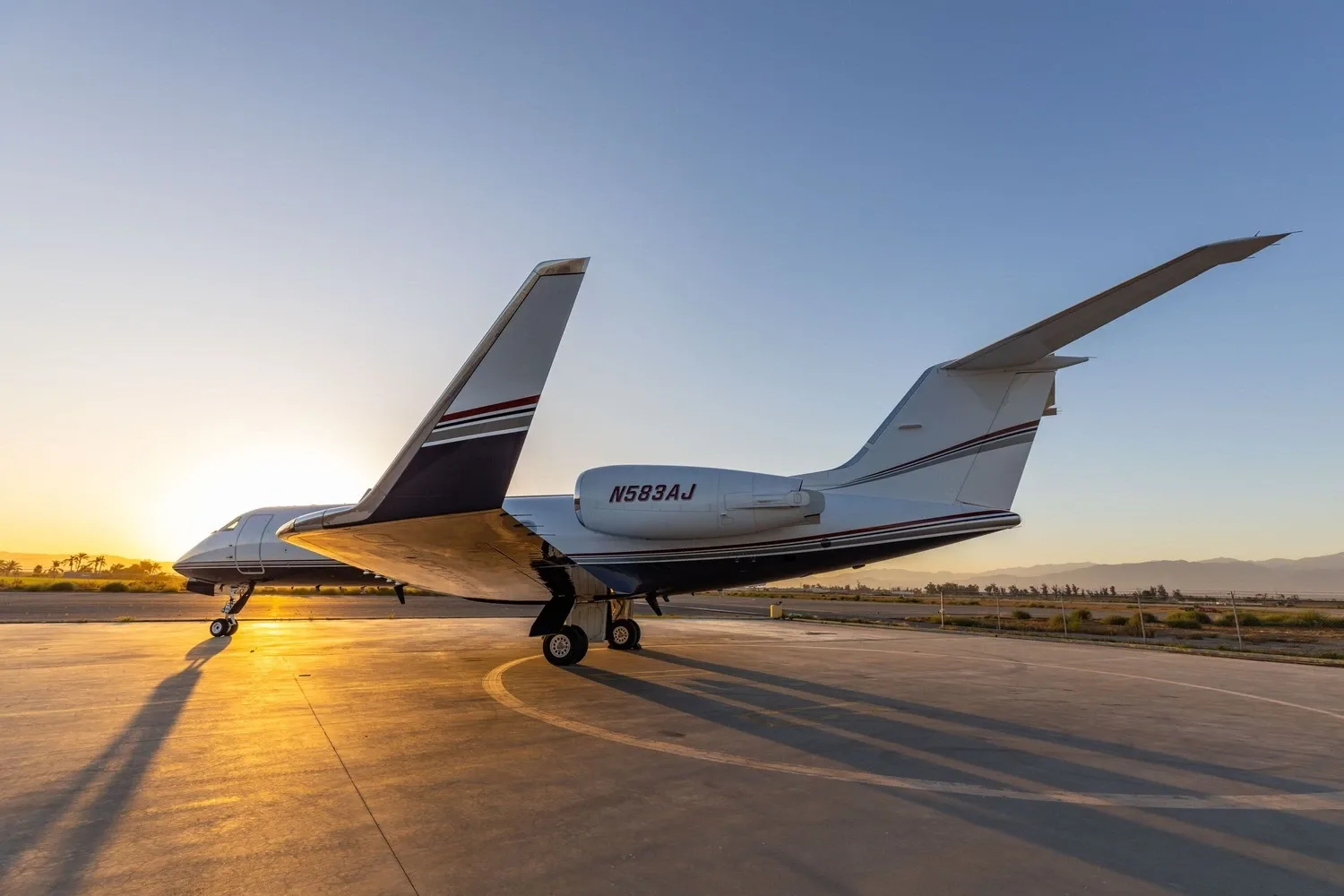 A white private jet with dark and red stripes on its tail and fuselage, parked on an airfield at sunset.