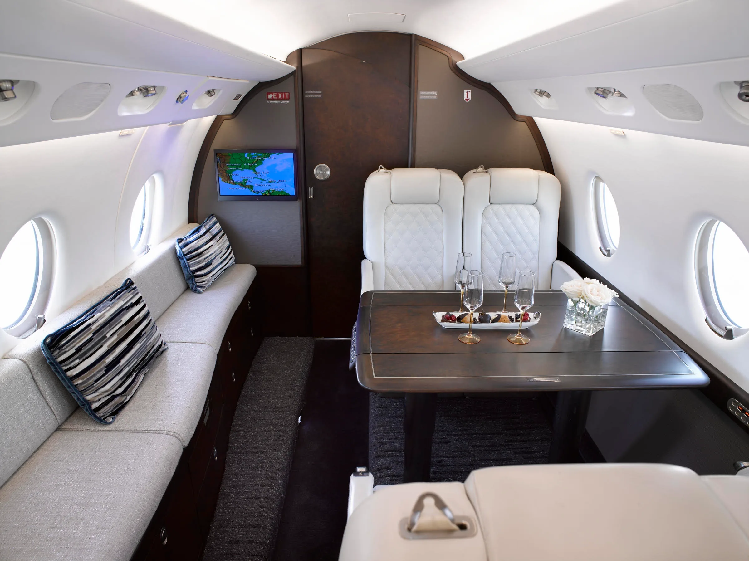 A wide shot of the private jet's cabin shows a comfortable lounge area with a sofa, a table with refreshments, and a screen displaying a map.