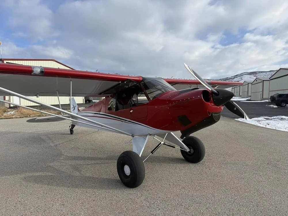 A red and white bush plane with large tires parked on an asphalt runway with hangars and snowy hills in the background.