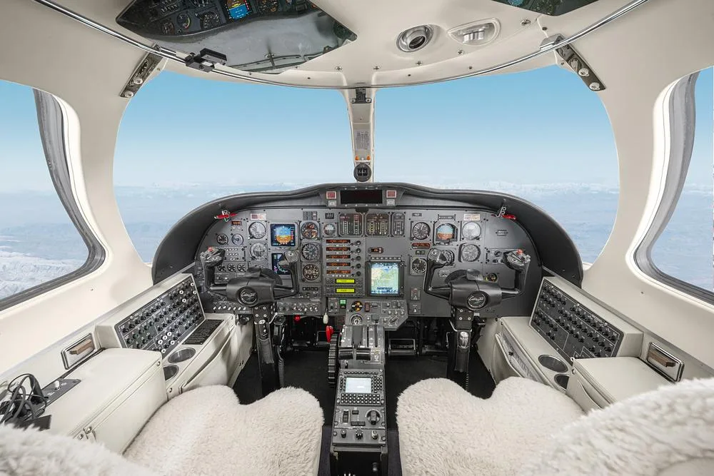 The cockpit of a private jet with a complex instrument panel, dual yokes, and a view of mountains from the windows.
