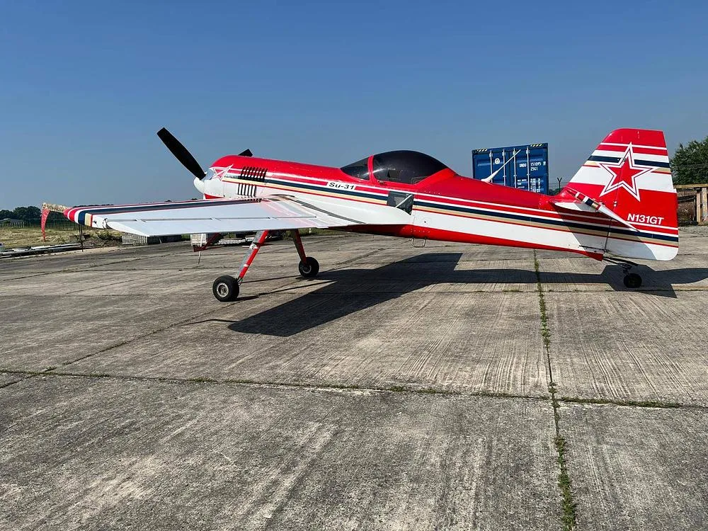 A red and white Sukhoi Su-31 aerobatic aircraft with registration N131GT on the tail is parked on a concrete tarmac, viewed from the side.