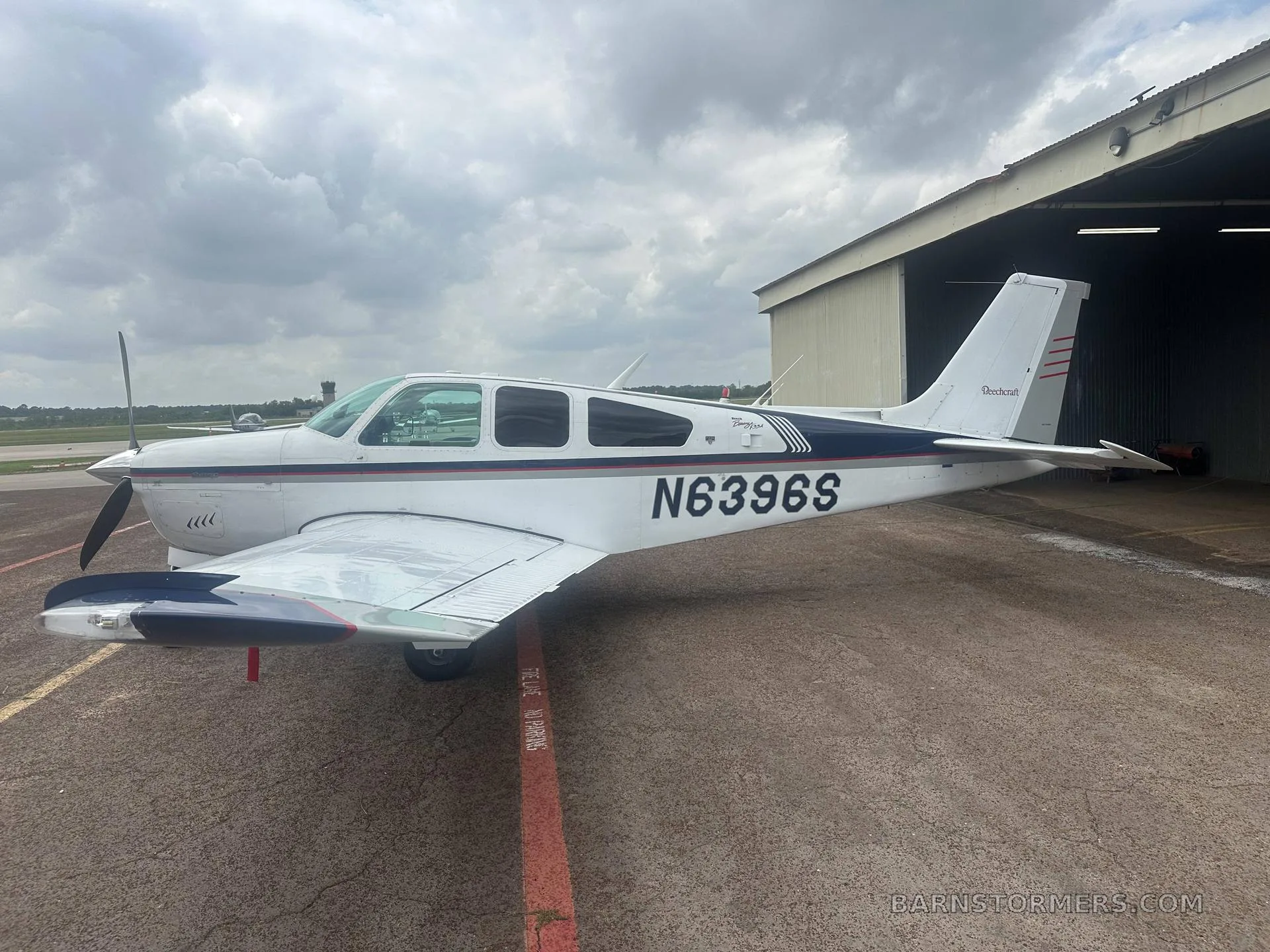 A white and blue Beechcraft Debonair N6396S parked on an asphalt apron next to a hangar under a cloudy sky, with an airport tower in the distance.