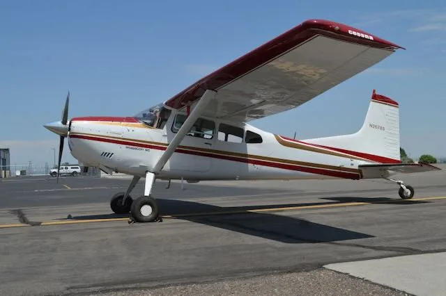A white Cessna Stationair 6 with red and gold stripes is parked on an asphalt tarmac under a clear blue sky.