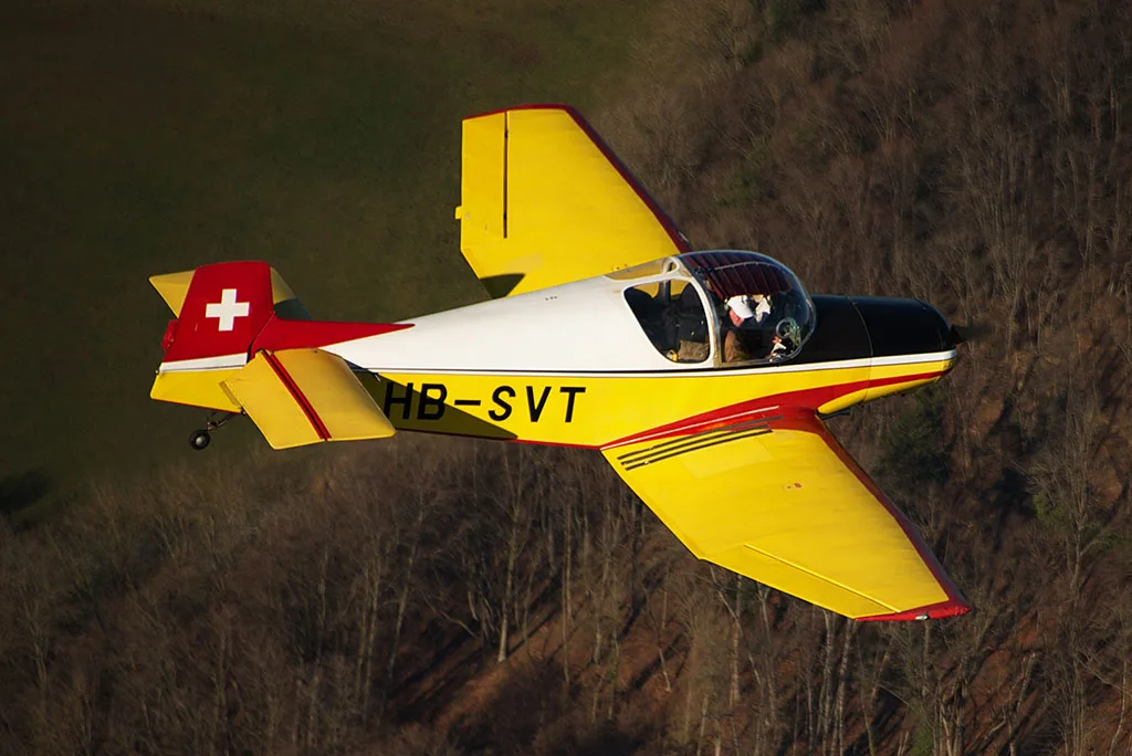 A yellow, red, and black single-engine aircraft (HB-SVT) flying over a brown, leafless forest.