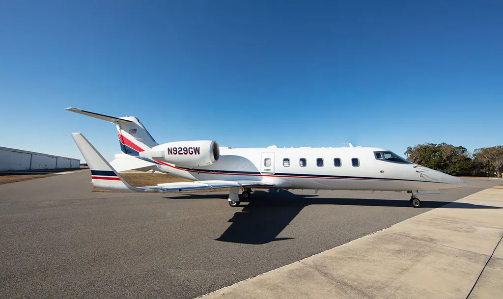 A white private jet with red and blue tail stripes, registration N929GW, parked on an airfield with a clear blue sky.