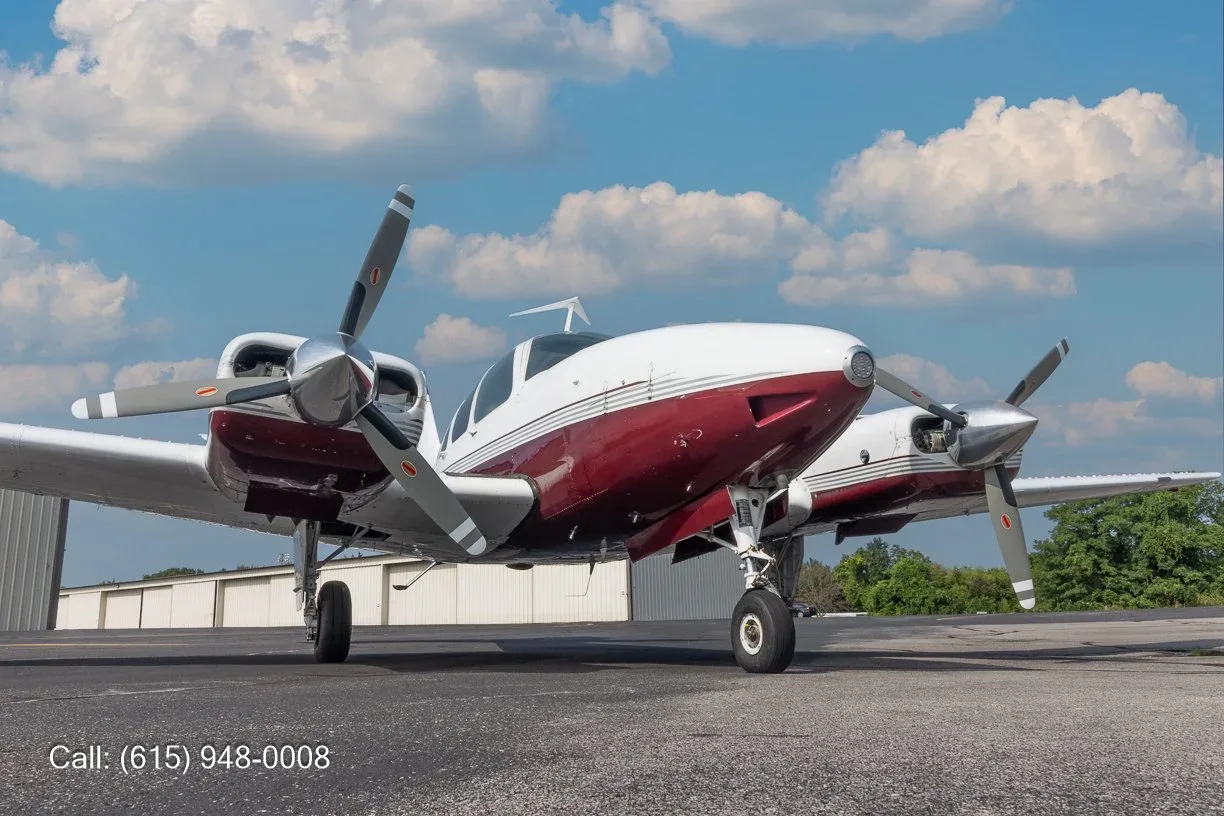 A white and maroon twin-engine propeller aircraft is parked on an airfield under a blue sky with white clouds.