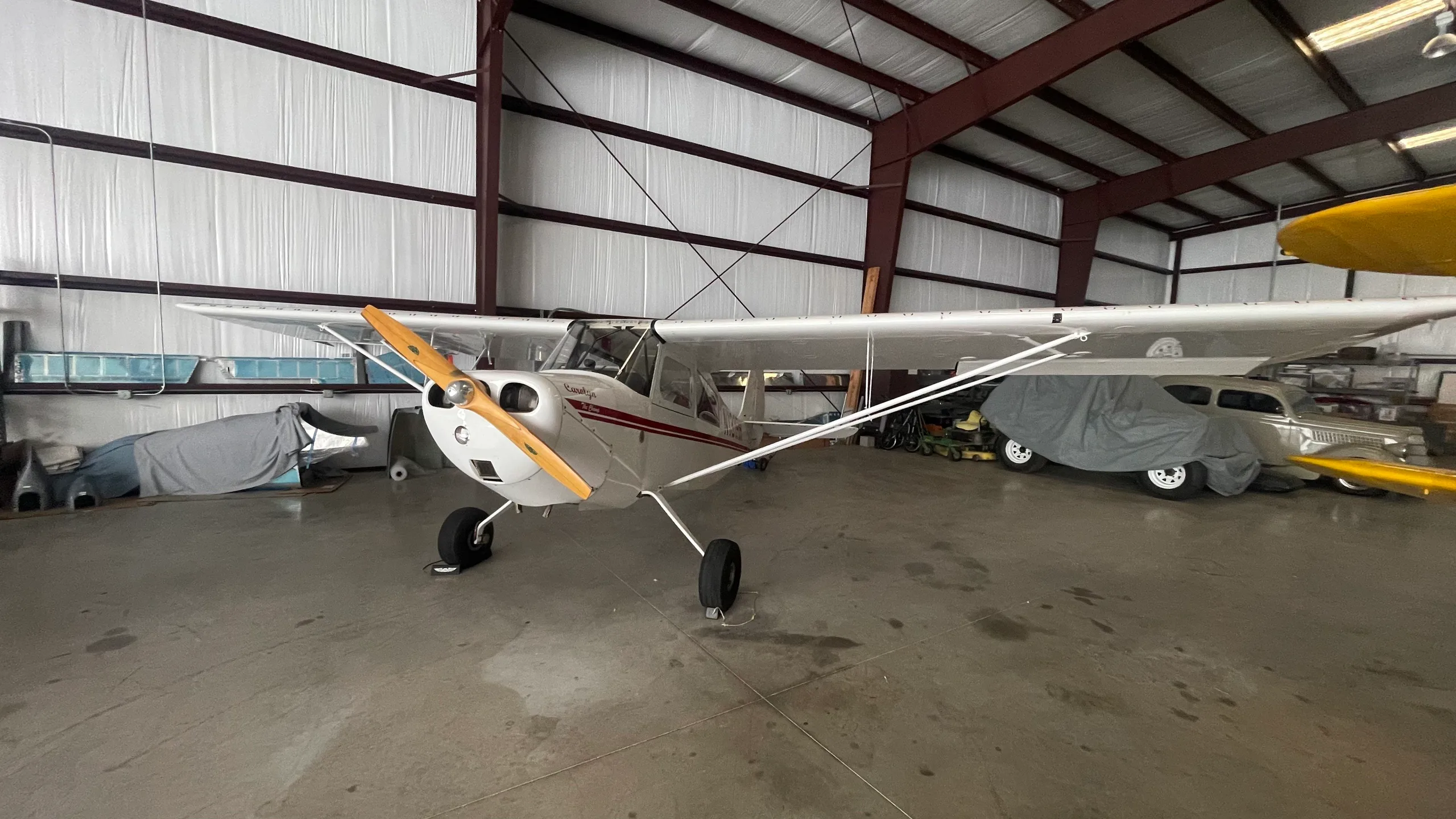 A white taildragger aircraft with red stripes and a wooden propeller is parked in a hangar, with other covered vehicles in the background.