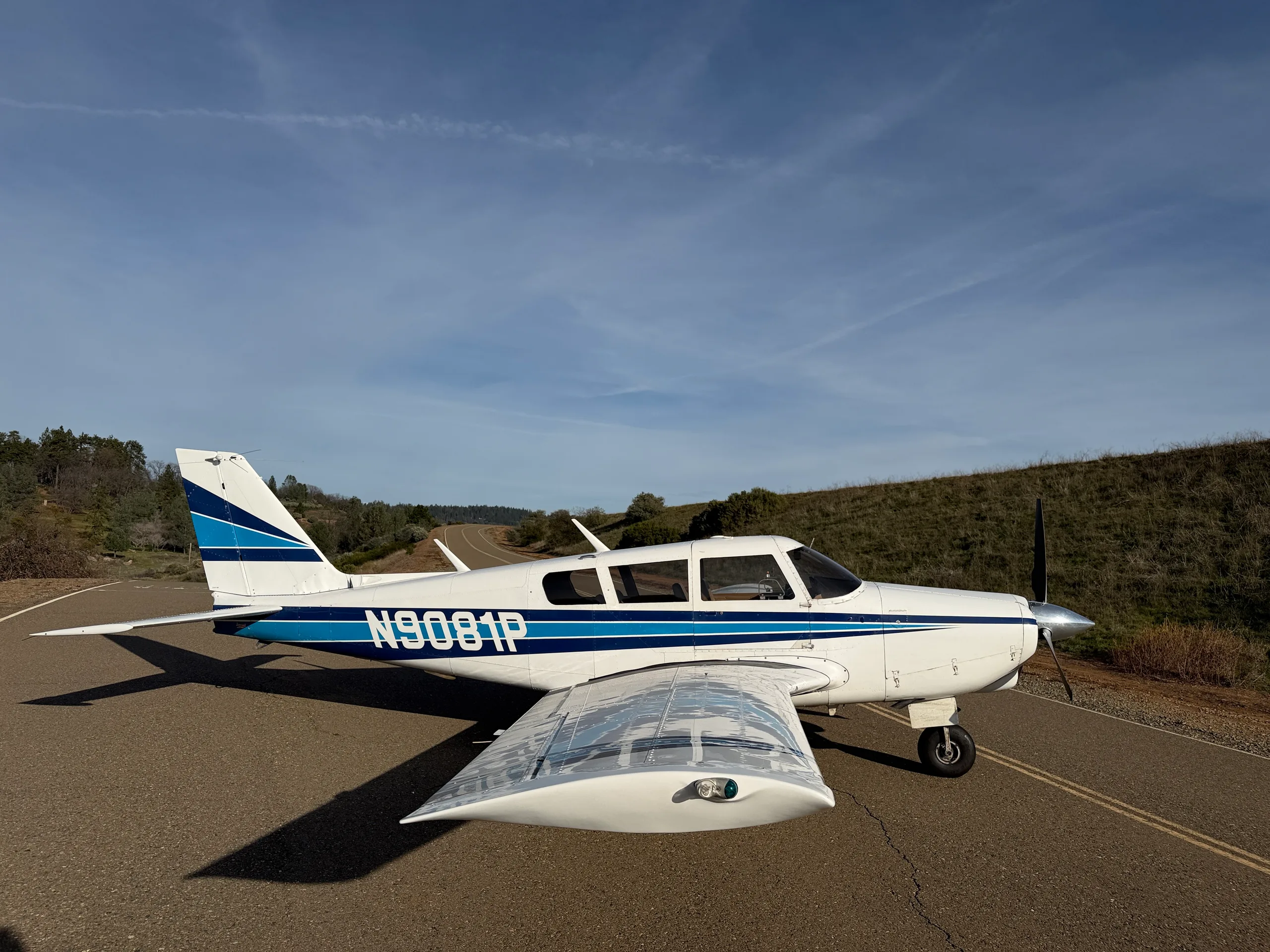 A white single-engine propeller aircraft with blue stripes (N9081P) is parked on an asphalt surface with hills and trees in the background.