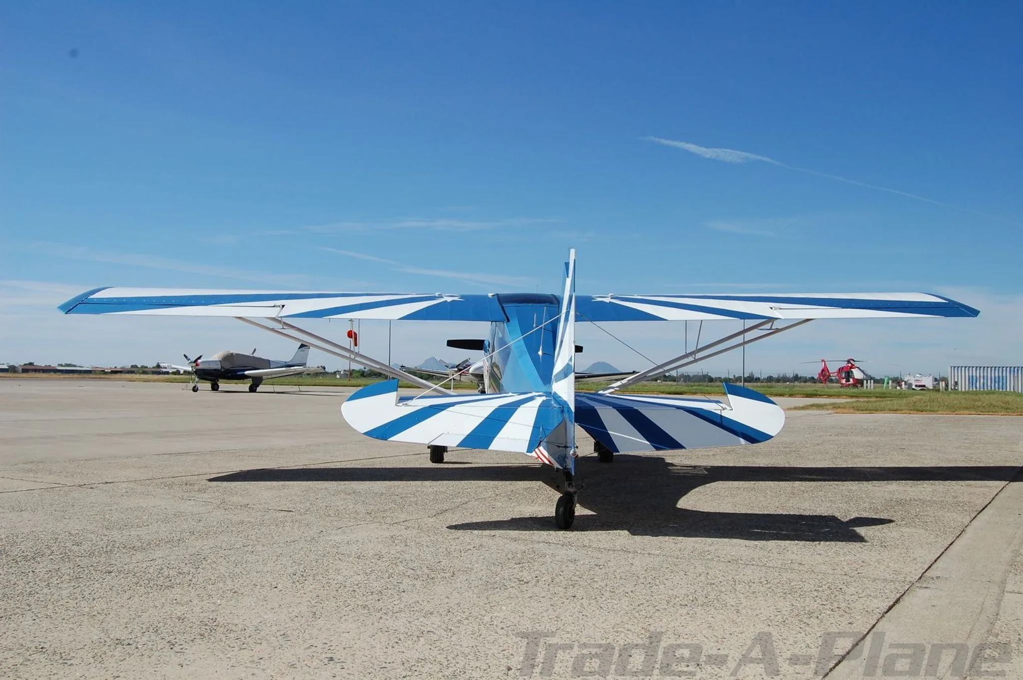 A rear view of the blue, white, and red single-engine aircraft, highlighting the striped tail and wings, parked on an airfield with other aircraft in the distance.