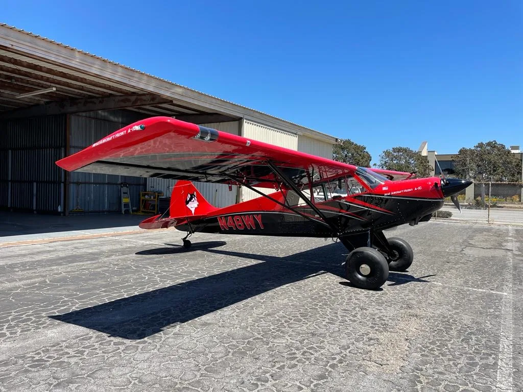 A red and black Aviat Husky A-1C aircraft with large tires is parked on an asphalt apron in front of a hangar on a sunny day.