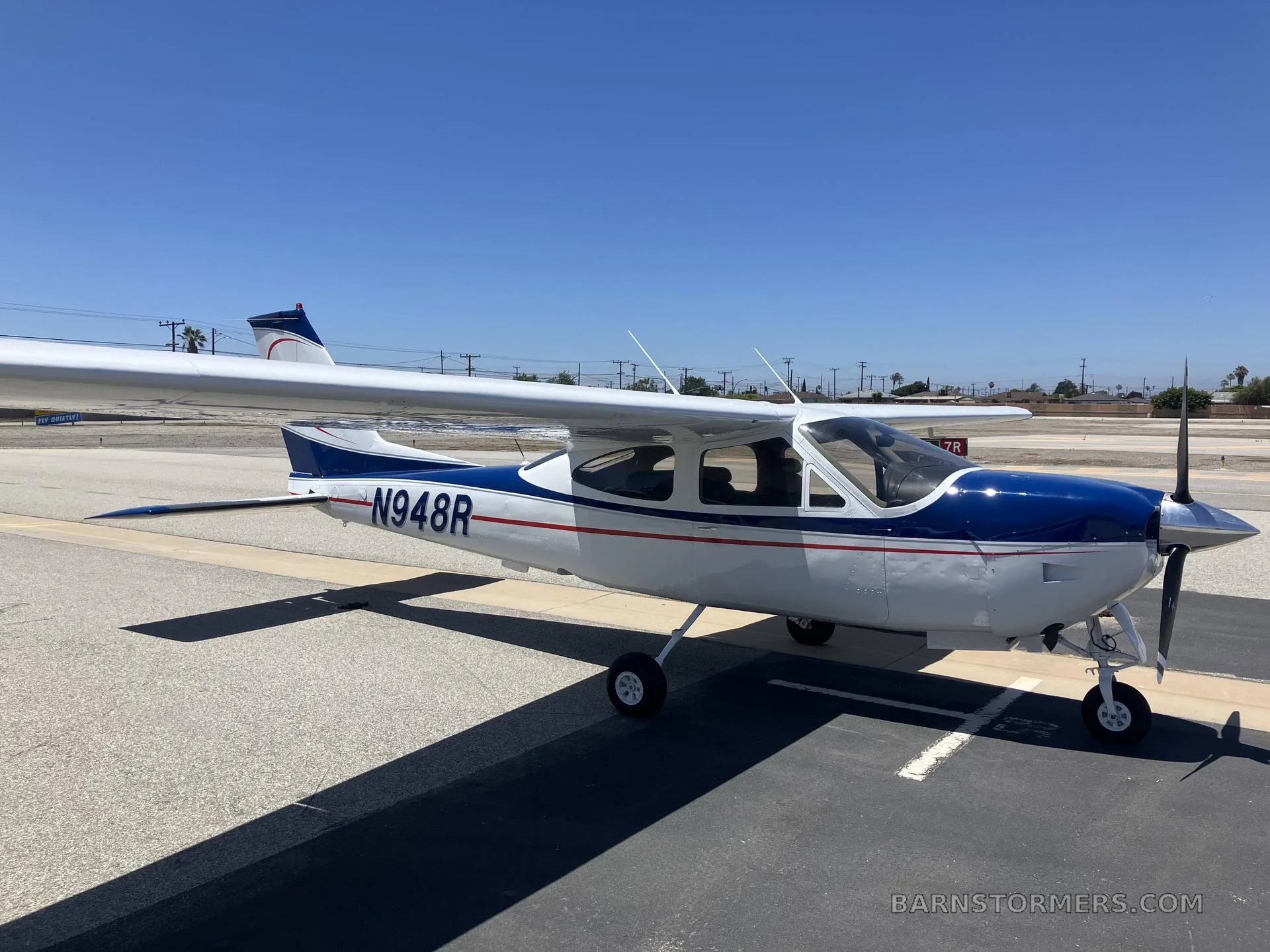 A white and blue Cessna 182 Skylane with registration N948R is parked on an asphalt ramp under a clear blue sky, viewed from the front-right side.