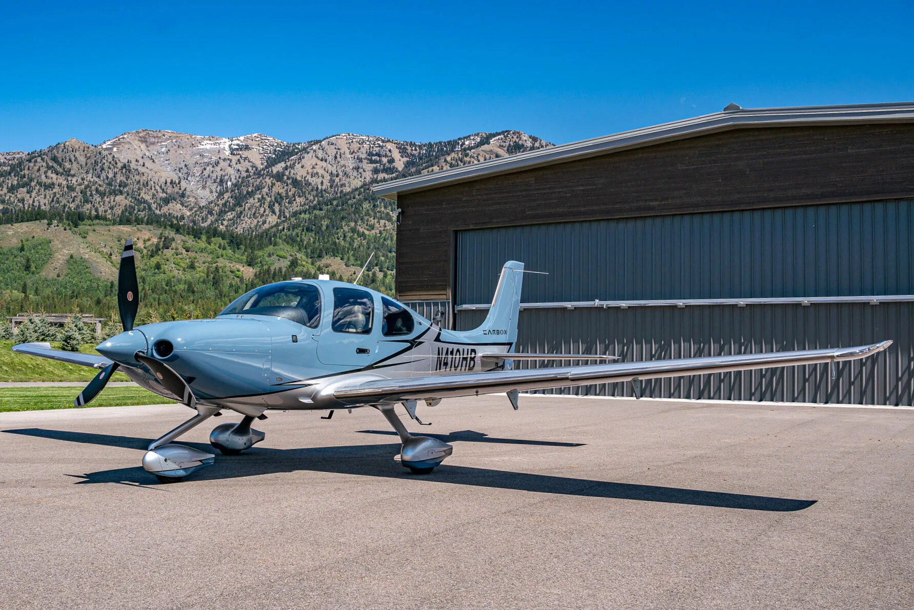 A light blue and silver Cirrus SR22T aircraft (N410HB) parked next to a hangar with mountains in the background.