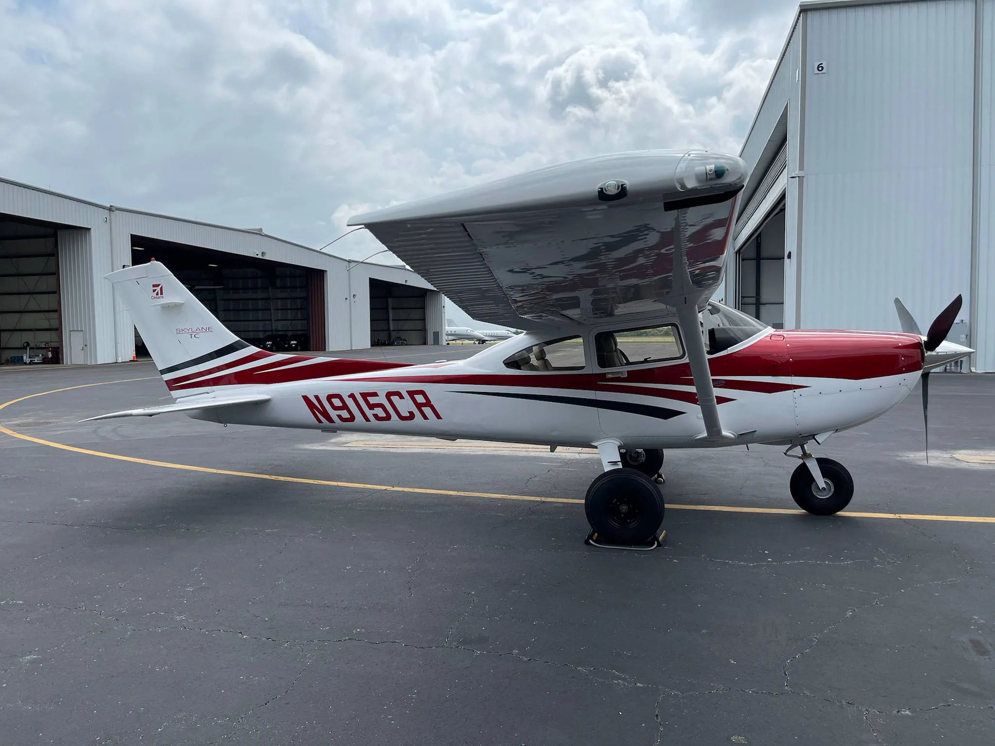A white Cessna Skylane TC (N915CR) with red and black stripes is parked on an airport tarmac with hangars in the background.