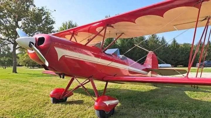 A red biplane with white stripes and wing accents, parked on a grassy field with trees in the background.
