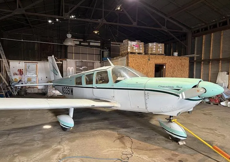A Piper Cherokee Six aircraft with white and light green paint scheme, parked inside a hangar.