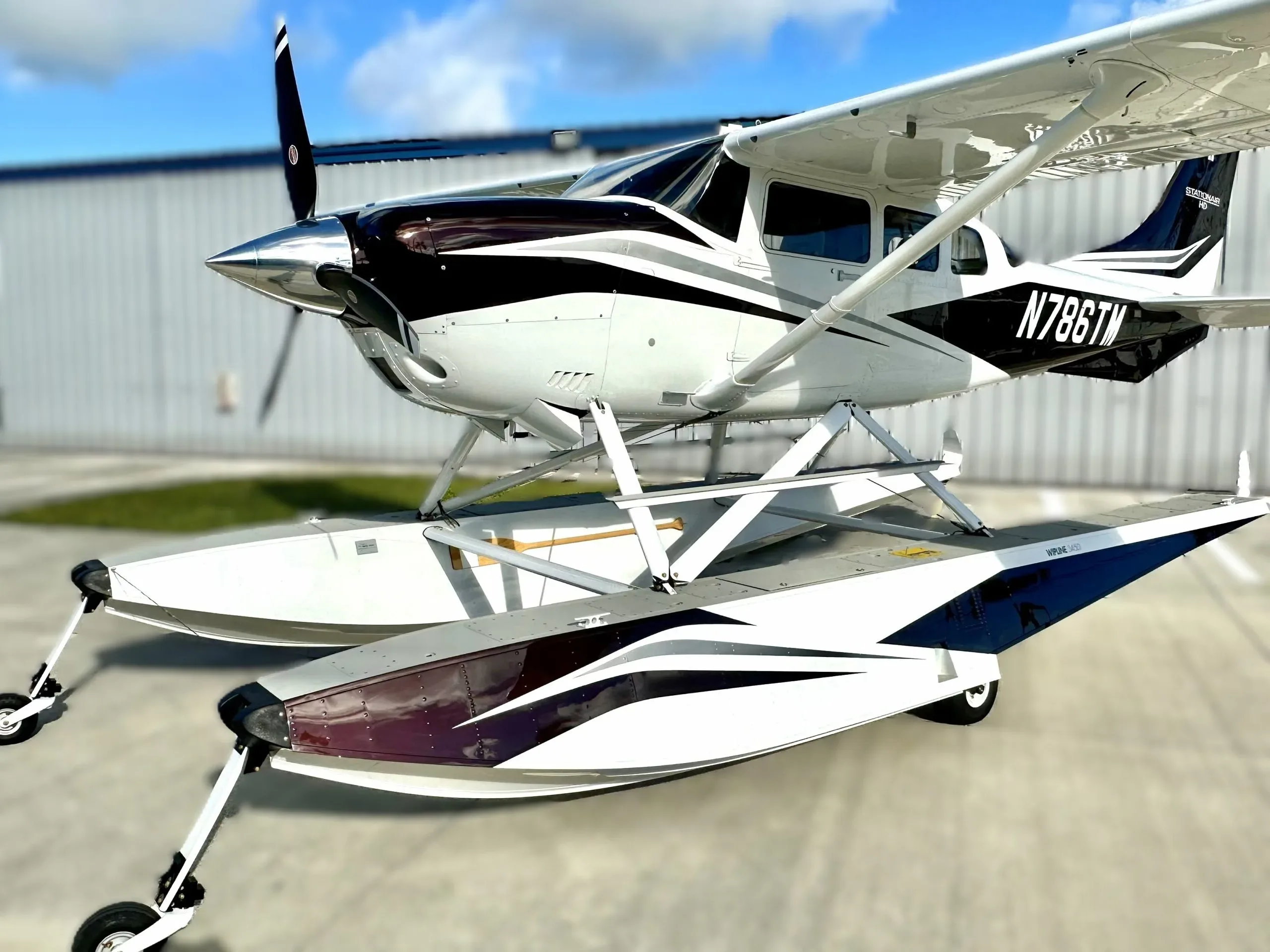 A white and dark brown Cessna Stationair HD seaplane with registration N786TM, parked on an airfield.