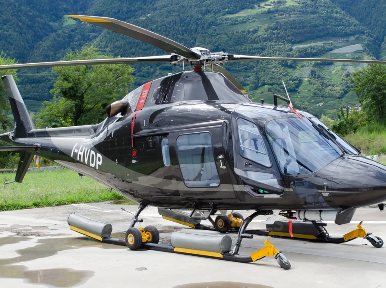 A black and grey AgustaWestland AW109 helicopter with registration I-HVDP is parked on a concrete pad with green mountains in the background.