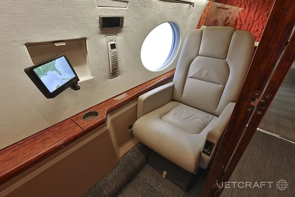 A close-up of a light beige leather cabin seat next to a window, with a small display screen mounted on the wall.