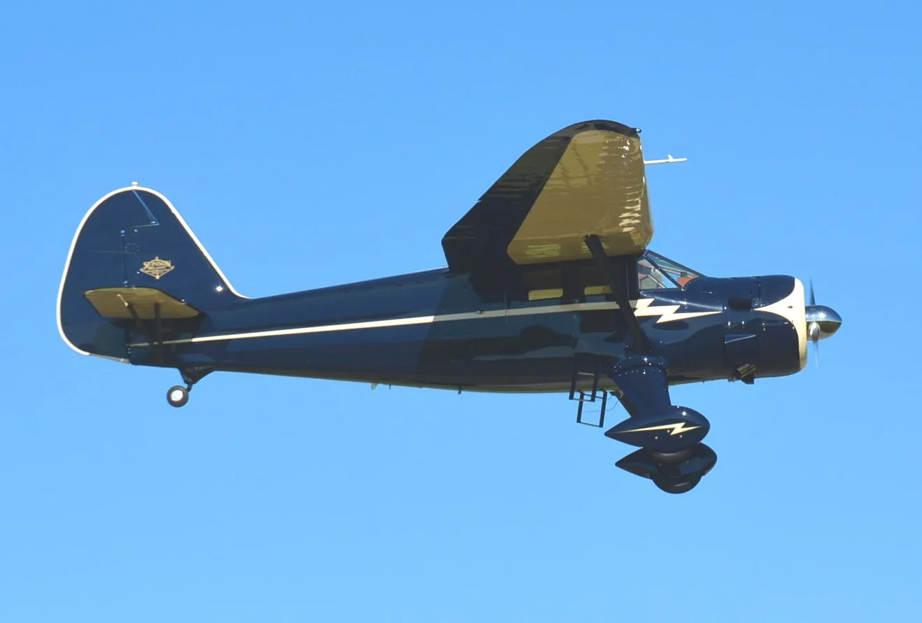 A dark blue Stinson Reliant aircraft with a white stripe and lightning bolt design, and a tan wing, flying against a clear blue sky.
