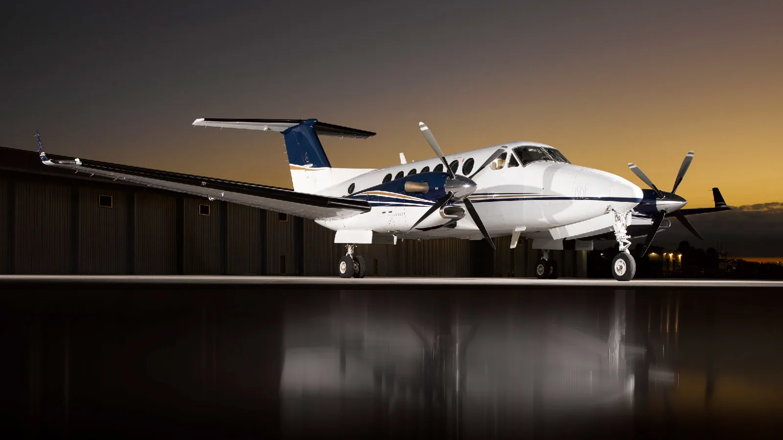 A white and blue twin-turboprop aircraft, likely a Beechcraft King Air, parked on a tarmac with a hangar in the background at sunset.
