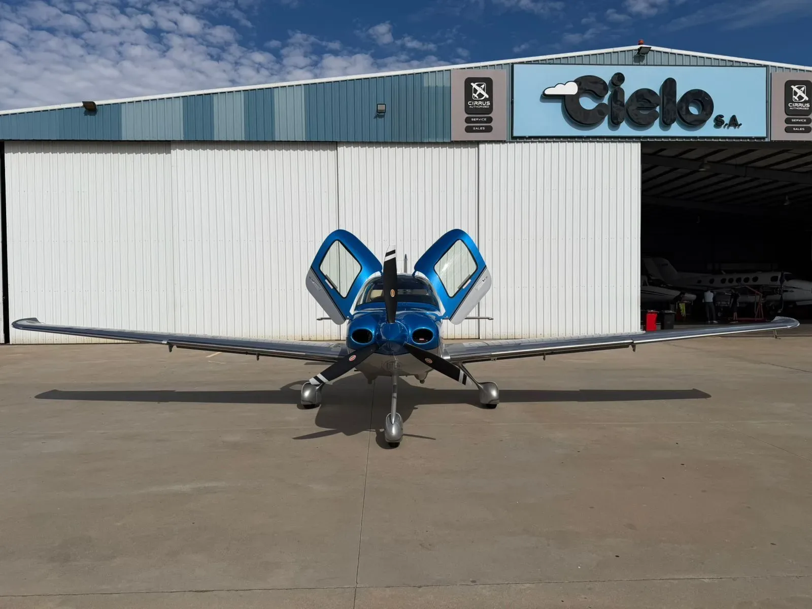 A blue and silver Cirrus SR22 aircraft with its gull-wing doors open, parked in front of a hangar.