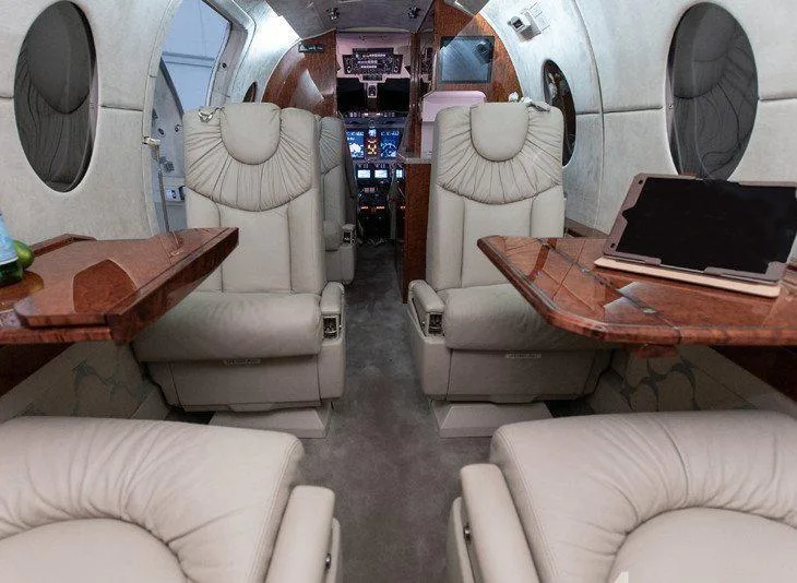 The interior cabin of a private jet, showing beige leather seats, wooden tables, and a view towards the cockpit.