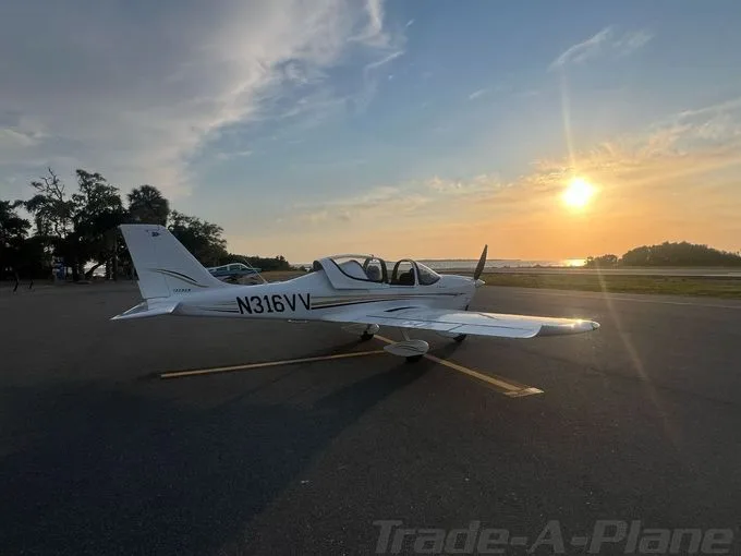 A white single-engine propeller aircraft, N316VV, is parked on an airfield at sunset with trees and water in the background.