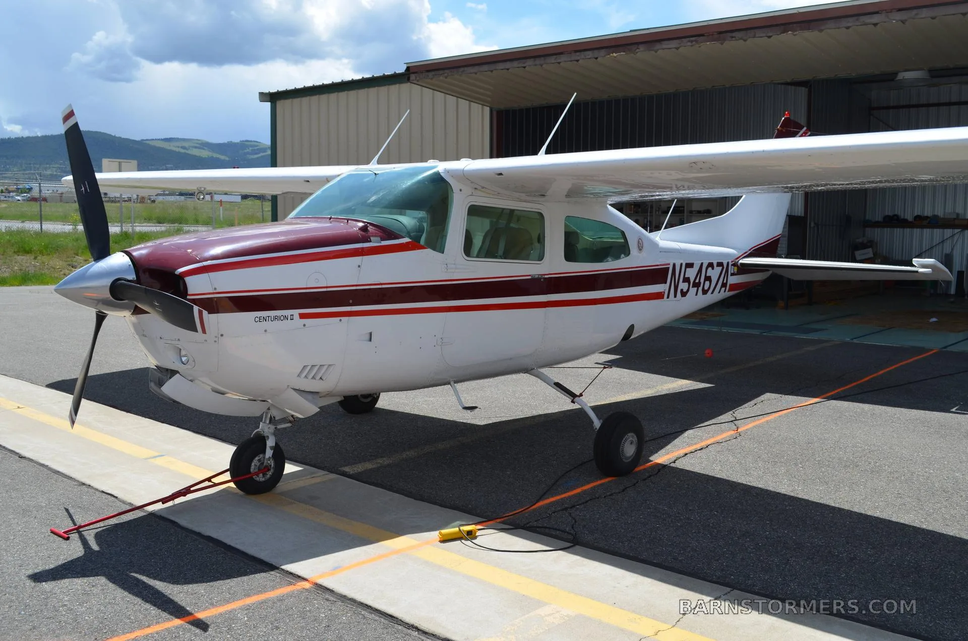 A white and maroon Cessna Centurion II (N5467A) parked on an asphalt ramp next to a hangar under a partly cloudy sky.