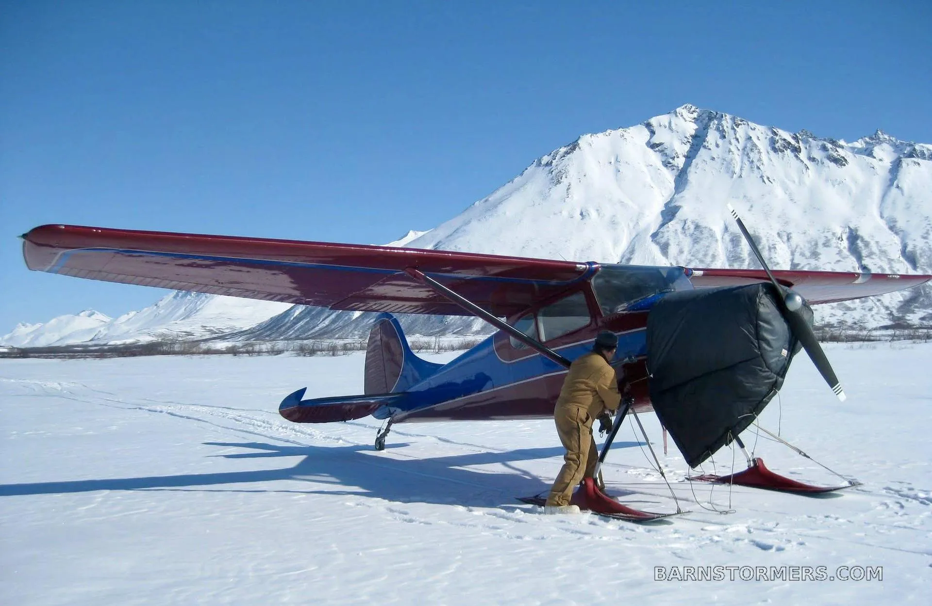 A red and blue high-wing aircraft on skis, with a black engine cover, parked in a snowy field with mountains in the background.